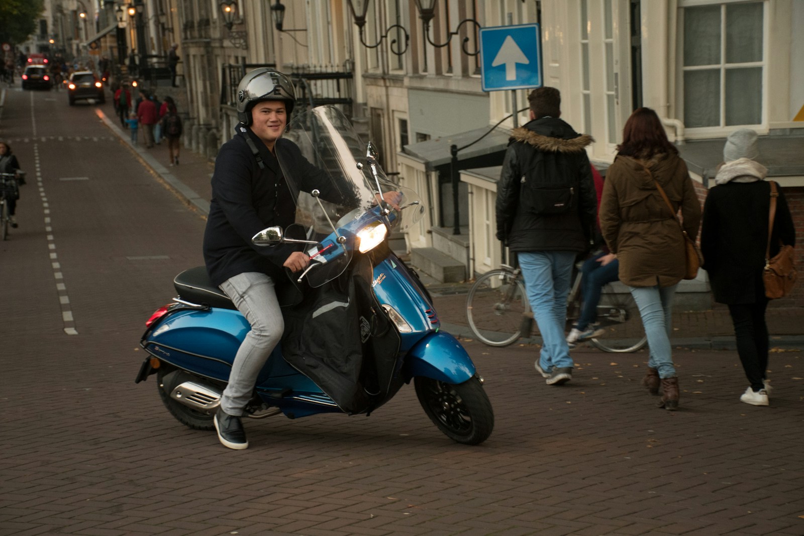 Persona con casco demi-jet alla guida di uno scooter blu su una strada lastricata